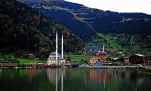 Mountains, green trees, the dome of a mosque, and a minaret, reflected in a lake and green plains on the lake’s bank