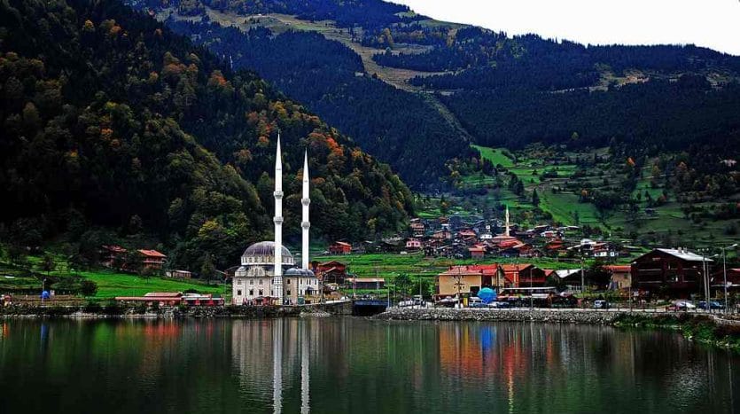 Mountains, green trees, the dome of a mosque, and a minaret, reflected in a lake and green plains on the lake’s bank