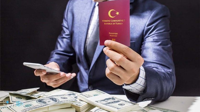 A person wearing a blue formal suit holds a Turkish passport in one hand and a mobile phone in the other hand In front of him are thousands of dollars on the table