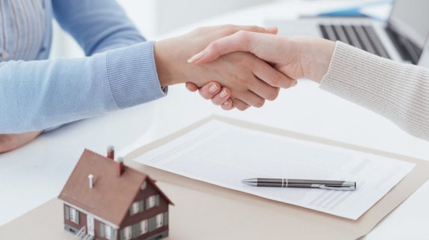 Two people shake hands on a desk containing work papers, a pen, and a small model of a house