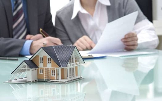 Two employees are sitting behind a desk, one of them is reading a white paper, and next to him is a model of a small house