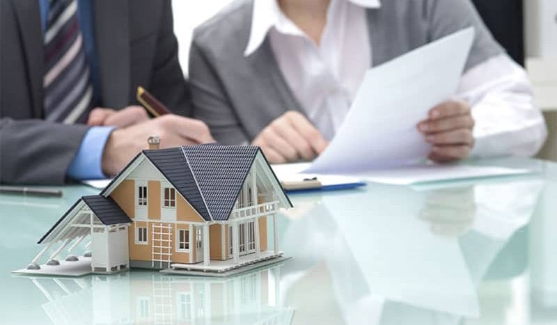 Two employees are sitting behind a desk, one of them is reading a white paper, and next to him is a model of a small house