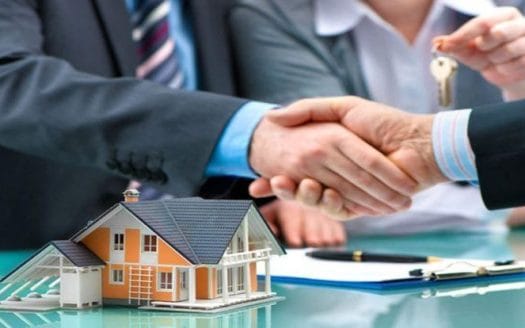 Two people shake hands and in front of them is a small model of a house, several papers, pens, and a house key. Real estate investment companies